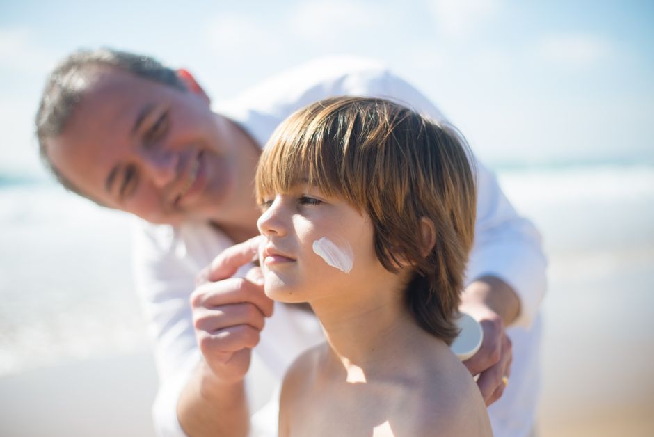 A father gently applies sunscreen to his son's face on a sunny day at the beach, ensuring 
