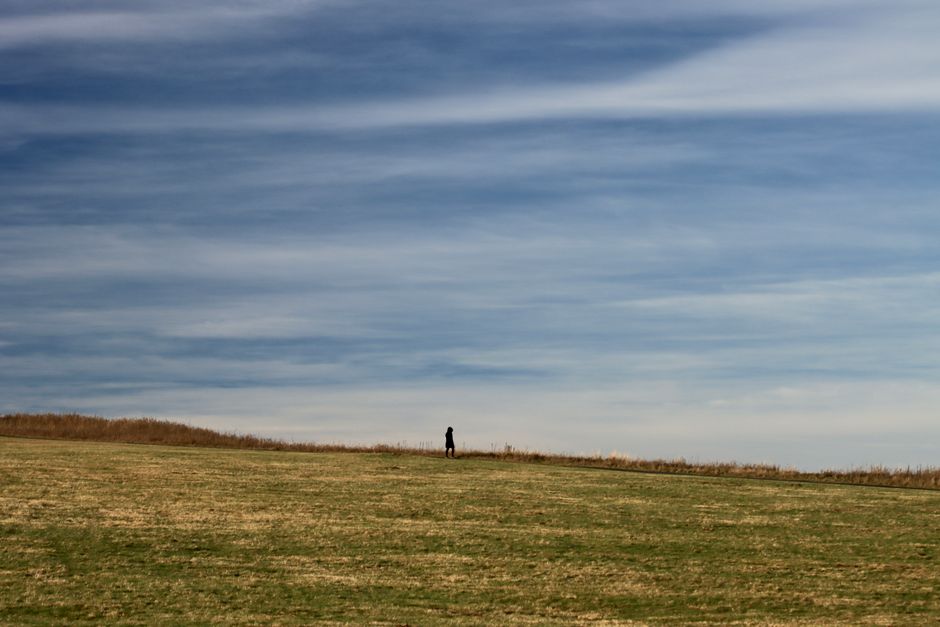 A lone person walks across an expansive grassy meadow beneath a cloudy sky, evoking solitu