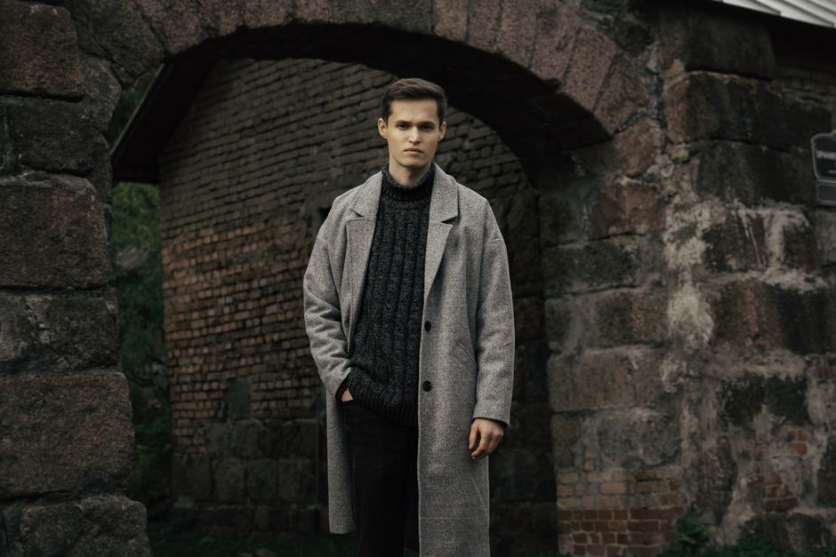 A man in a gray coat standing by a historic brick and stone archway in Saint Petersburg, R