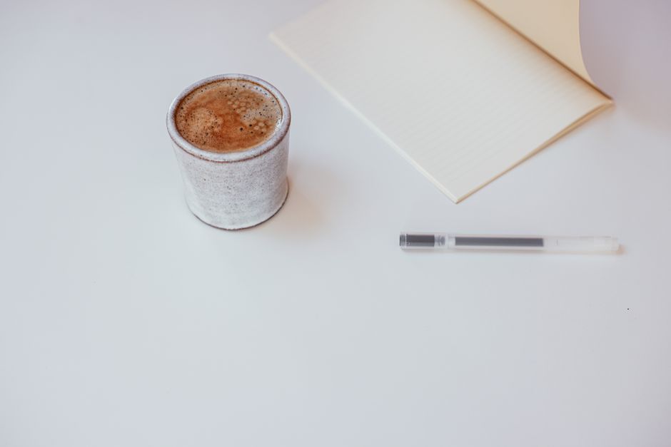 A minimalist workspace setup featuring a cup of coffee, notebook, and pen on a white surfa