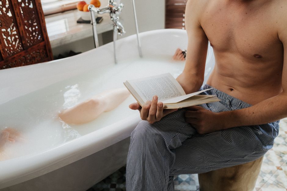 A serene indoor scene featuring a topless man reading while sitting beside a bathtub.