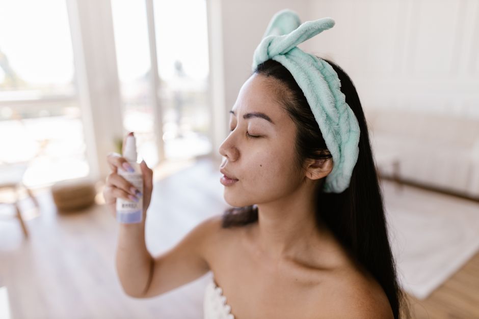A woman enjoying a home spa treatment, spraying facial mist in a relaxed indoor setting.