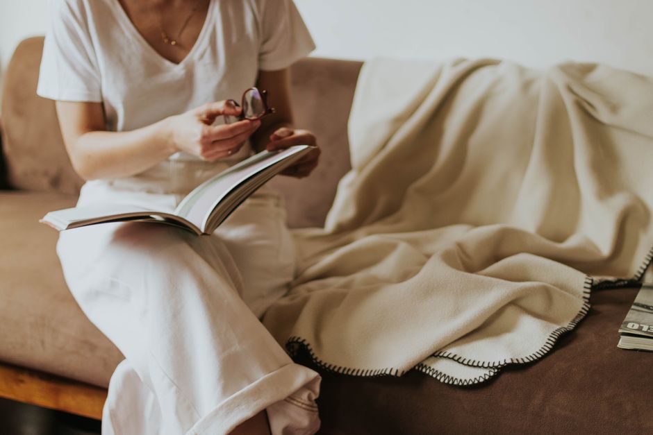 A woman sitting on a sofa, reading a book and holding eyeglasses, conveying relaxation ind
