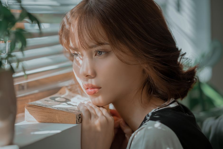 A young woman gazing thoughtfully out of a window with soft, natural light streaming in.