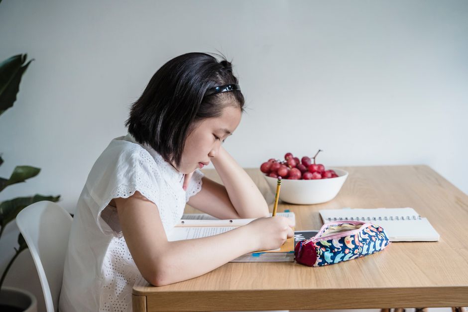 Child focuses on homework at a wooden table indoors, surrounded by study materials.