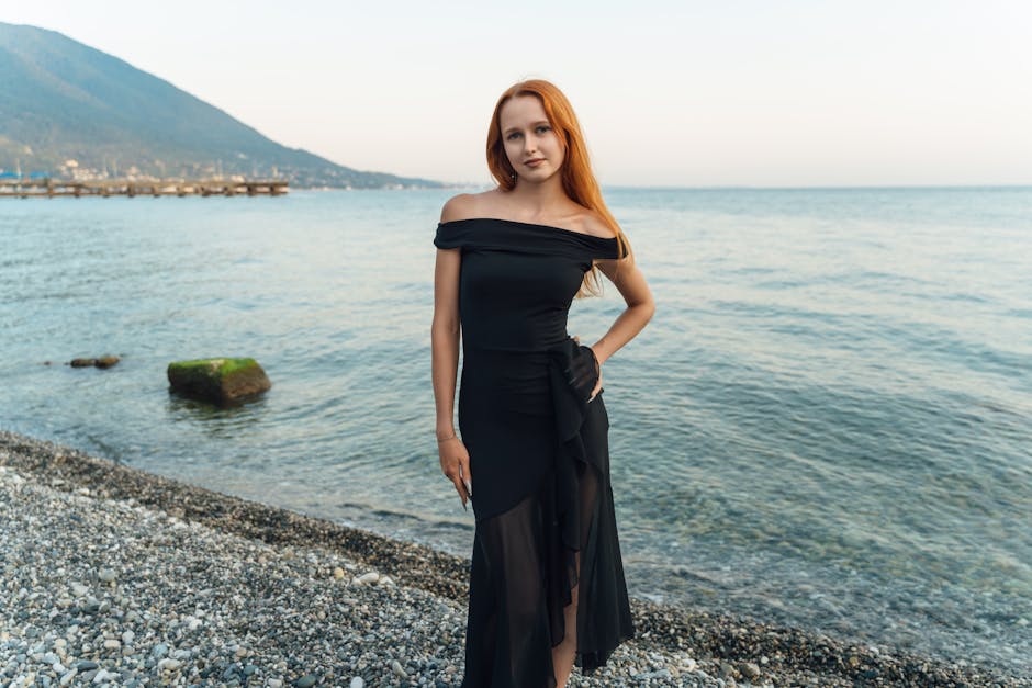 A stylish woman in a black dress poses by the picturesque sea shore in Гагра, perfect for travel and fashion themes.