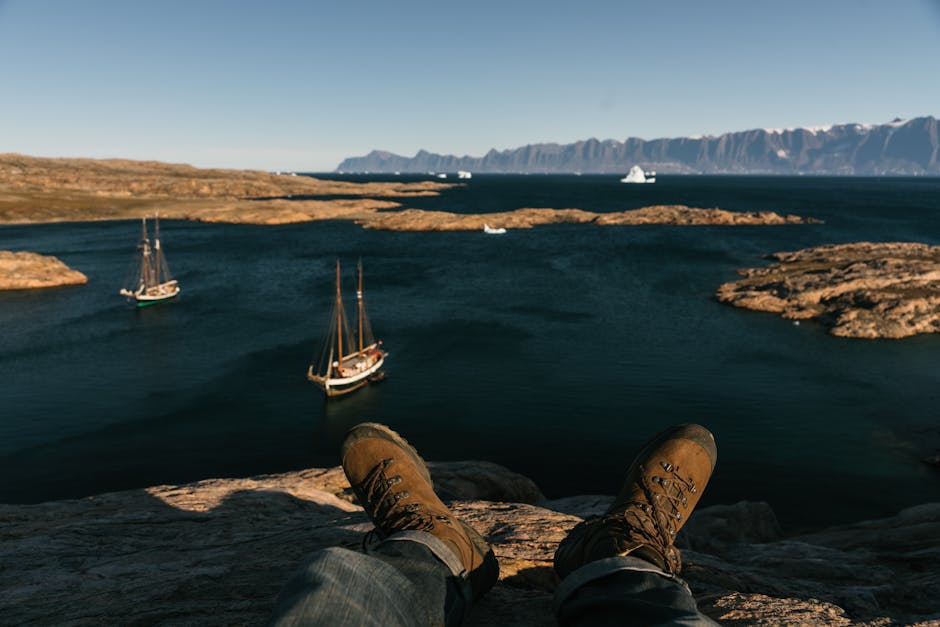 Person sits on rocky shore with sailboats and cliffs in view, ideal for outdoor adventure enthusiasts.
