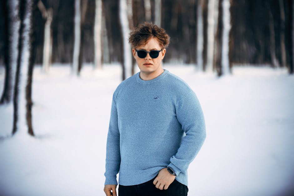 A young man wearing a blue sweater and sunglasses in a snowy forest setting.