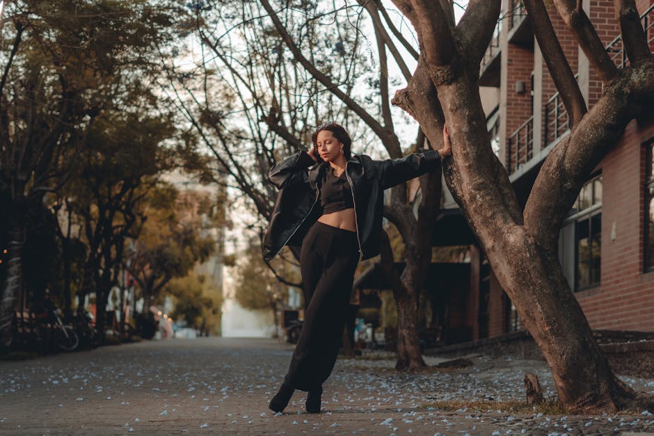 Woman in black outfit leans against tree in urban street, exuding elegance.