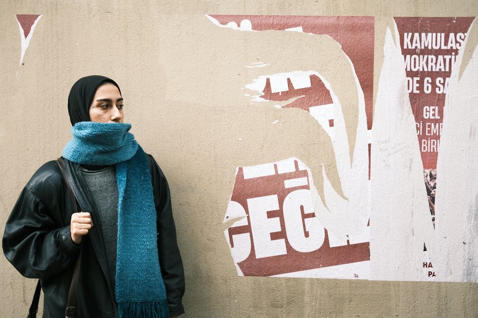 Woman in hijab with leather jacket and blue scarf standing by urban wall.