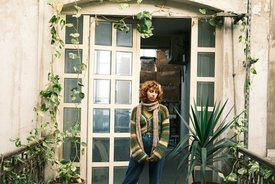 Woman in cozy attire stands in doorway with trailing plants in a rustic apartment setting.