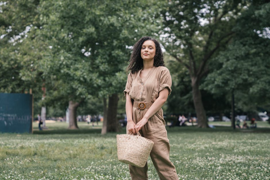 Fashionable woman in a beige jumpsuit standing in a lush green park holding a straw bag.