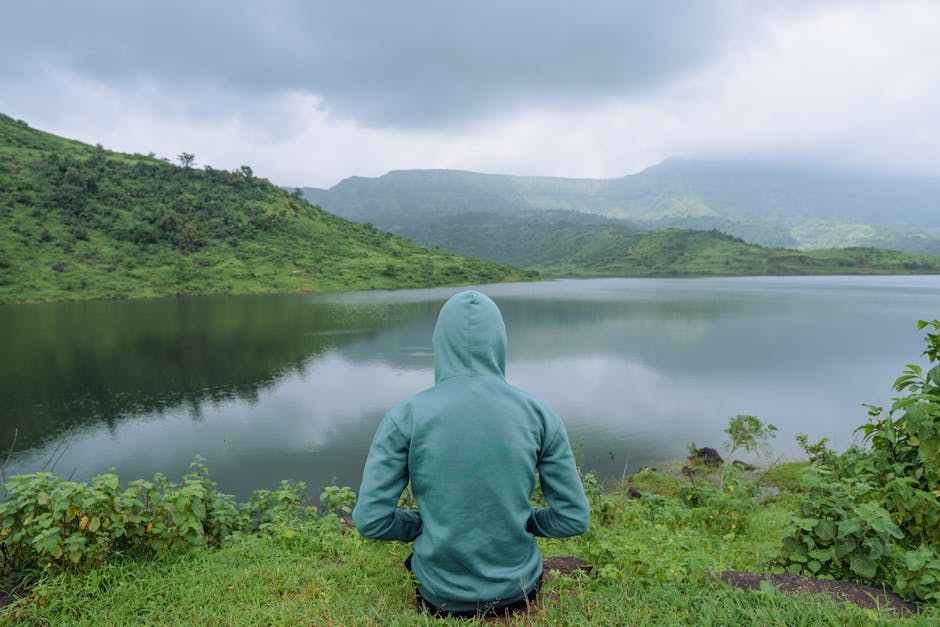 Person in teal hoodie sits by a tranquil lake in Dhom, India, surrounded by lush green hills.