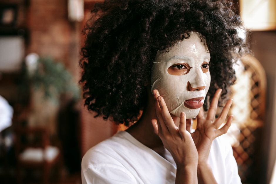 Calm young African American lady with curly hair applying moisturizing sheet mask while re