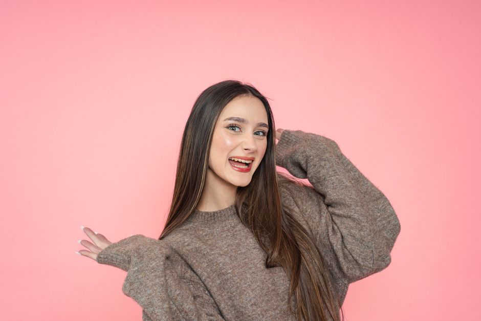 Cheerful young woman in a brown sweater poses against a pink backdrop, expressing joy and 