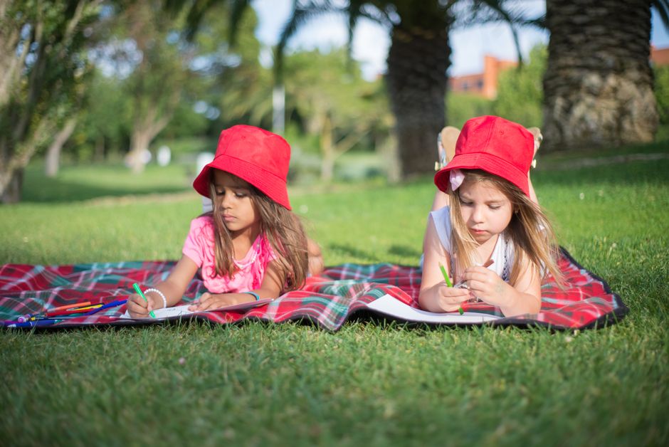 Children lying on a blanket, drawing in a park with red hats and sunny weather.