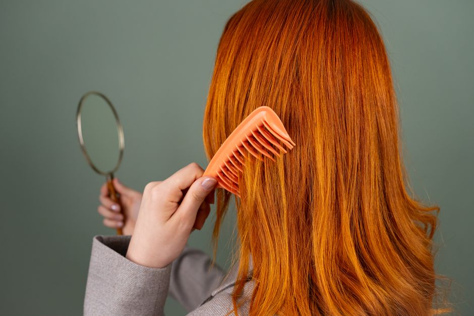 Close-up of a woman with red hair combing while holding a mirror.