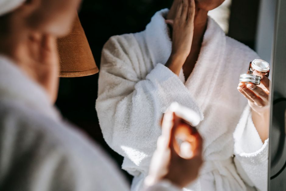 Crop anonymous female in white bathrobe applying moisturizing facial cream on face while s