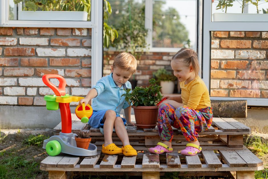 Two children sitting on wooden pallets, playing with toys, and watering plants. Bright and joyful outdoor scene.