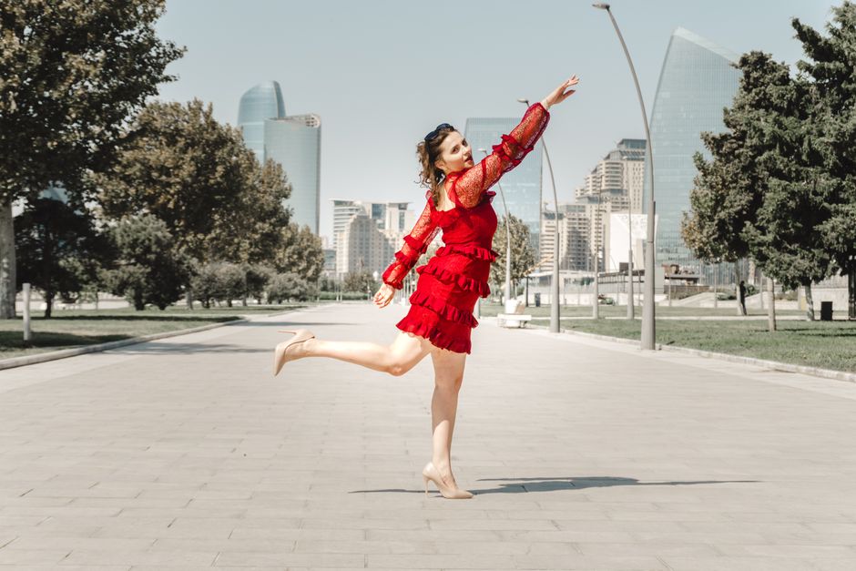 A woman in a vibrant red dress leaps gracefully on a paved pathway with modern city buildings in the background.