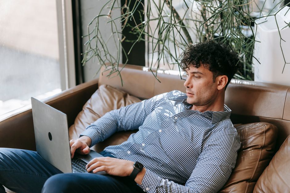 A man in smart casual attire works on a laptop while sitting on a sofa indoors.