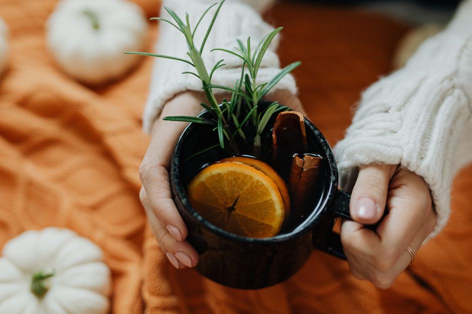 Hands holding a warm cup with spices and orange slices, surrounded by autumn decorations.