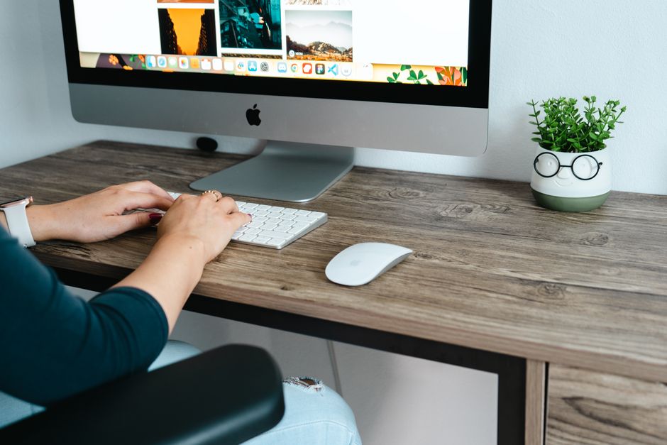 A woman's hands typing on a keyboard at a wooden desk with a computer setup.