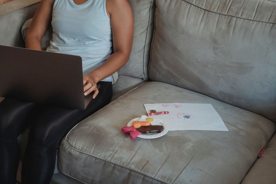 Crop anonymous female in casual wear sitting on comfortable sofa with toy sweets and drawing and browsing internet on laptop
