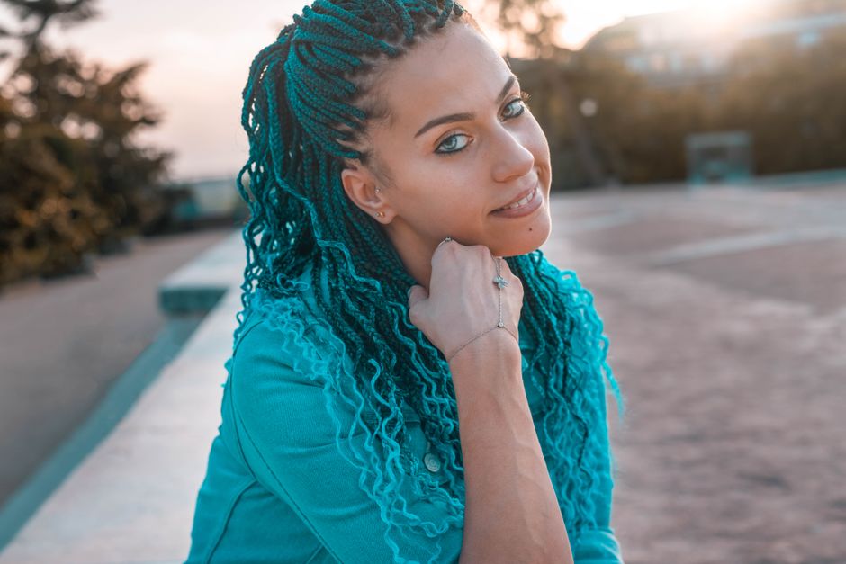Portrait of a stylish woman with braided blue hair sitting outdoors, enjoying a sunny day.