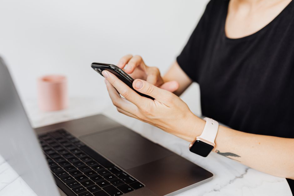Close-up of a woman using a smartphone with a laptop on an office desk, showcasing technology in the workplace.