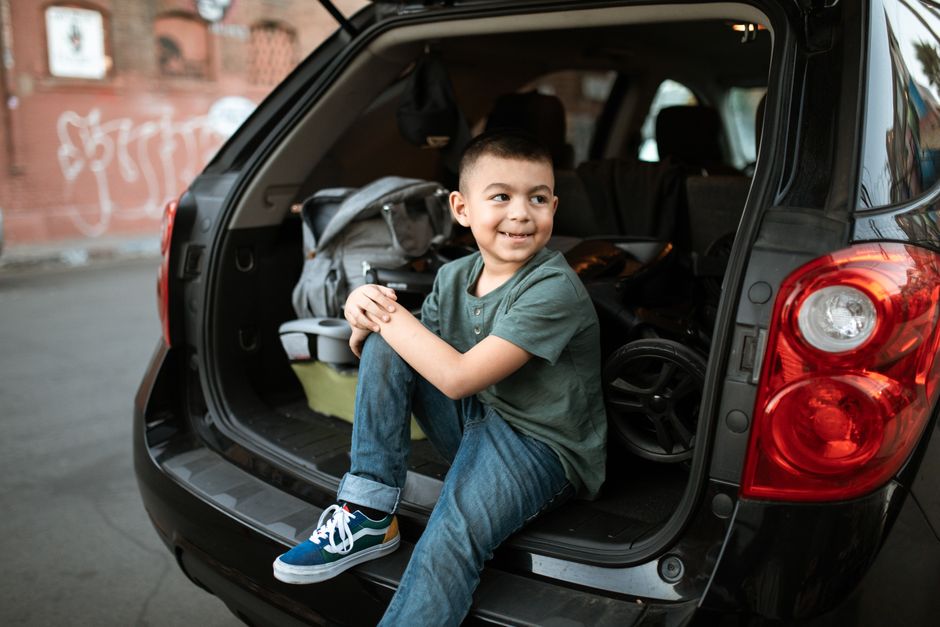Smiling child sitting in the trunk of an SUV on an urban street, wearing casual clothes.
