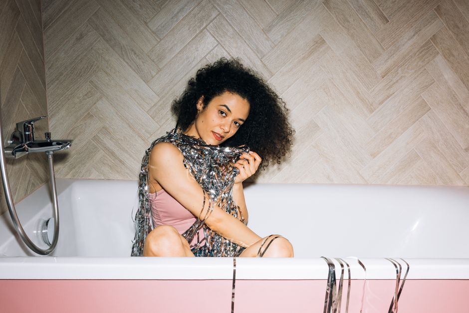Stylish young woman with afro hair posing playfully in a bathtub, wrapped in shiny streame