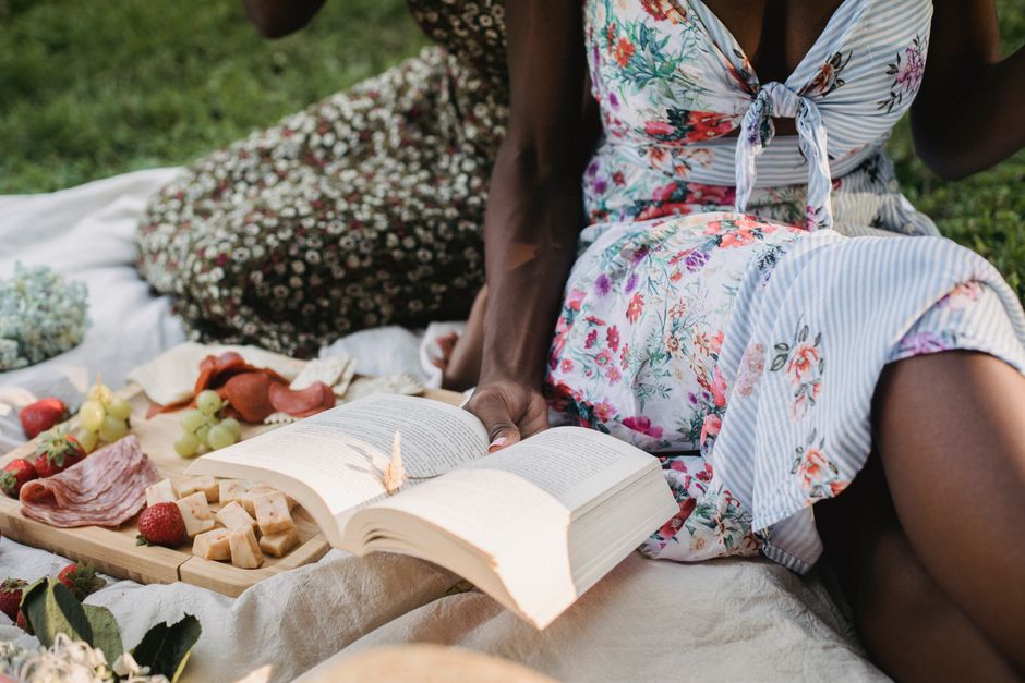Two friends enjoying a picnic with food and books on a sunny day, sitting on a blanket in the park.
