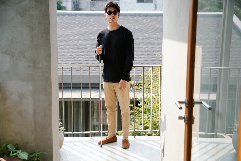 Blind man with a walking stick standing in a doorway wearing casual clothes and sunglasses.