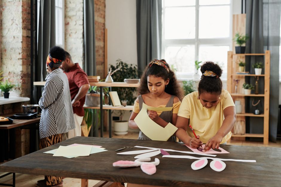Two children crafting with paper indoors while adults cook in the background.