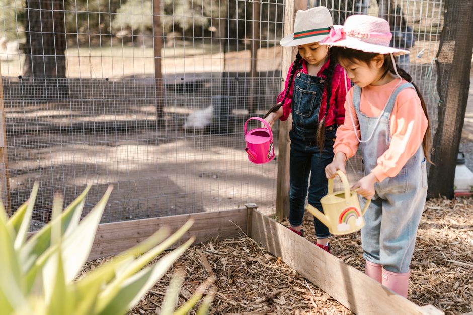 Two young girls gardening outdoors with watering cans and sun hats, fostering nature appre