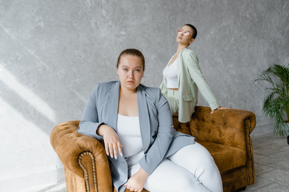 Two confident women posing in chic blazers on a vintage brown couch in a modern setting.