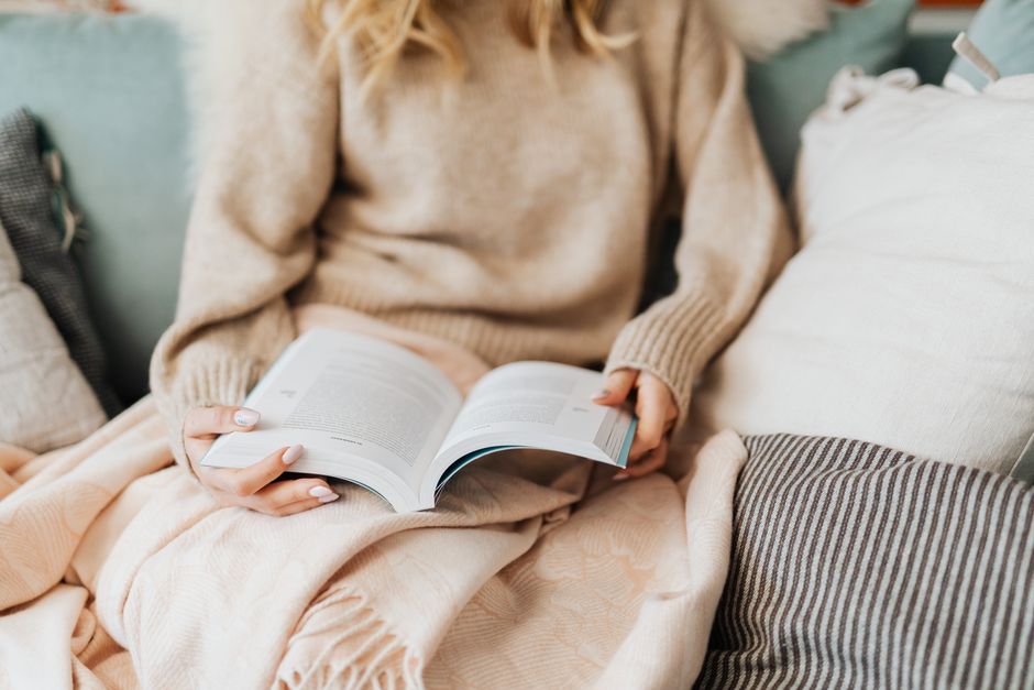 A person with manicured nails enjoys a book while wrapped in a cozy blanket.