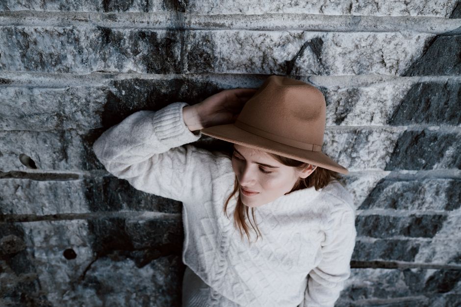Woman wearing a fedora and white sweater, leaning against a textured stone wall for a casu
