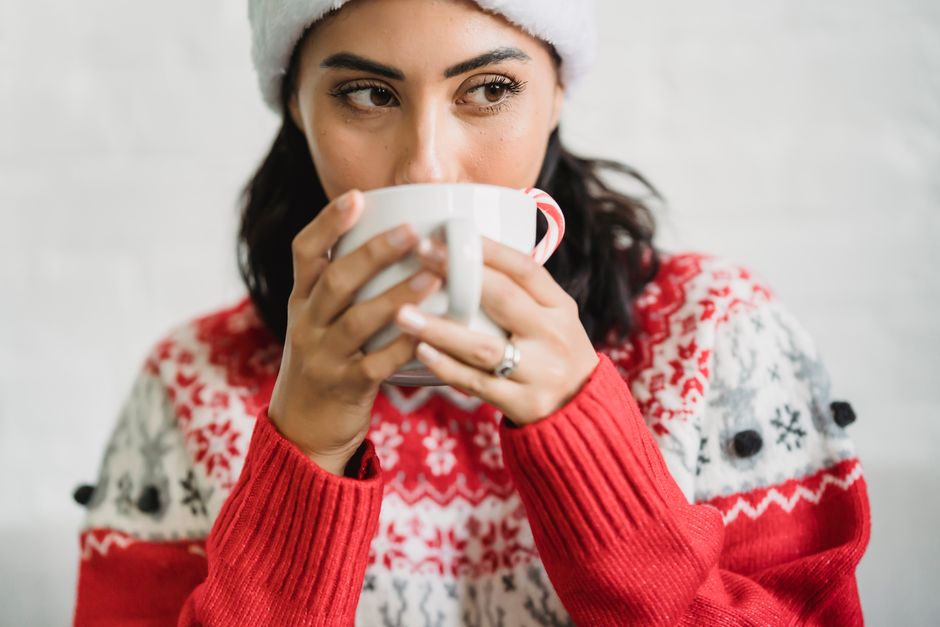 Young lady in Santa hat and sweater enjoying cup of cacao with candy cane in light room an